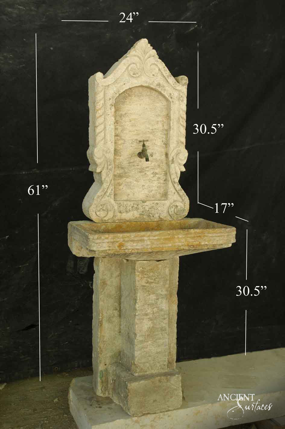 Tall cream limestone pedestal sink with a shallow moulded basin and a carved arched backplate with scrolled crest and small spout